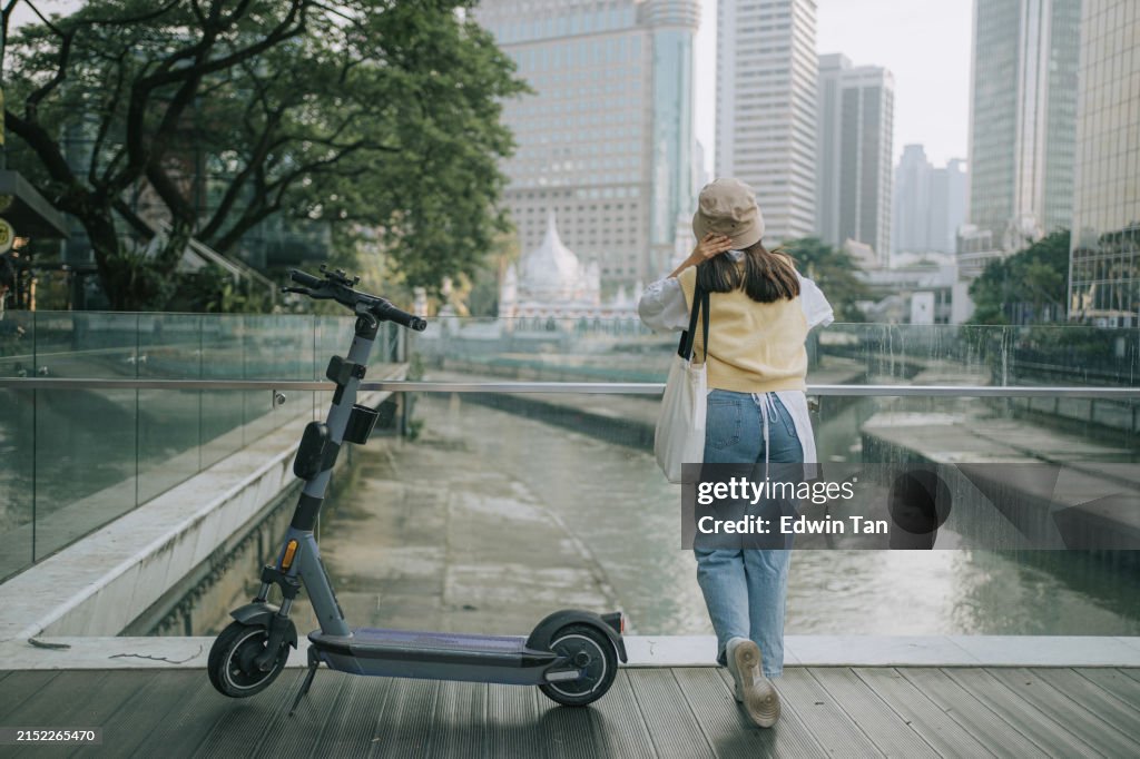 Rear view young female tourist looking at Kuala Lumpur cityscape leaning beside electric push scooter