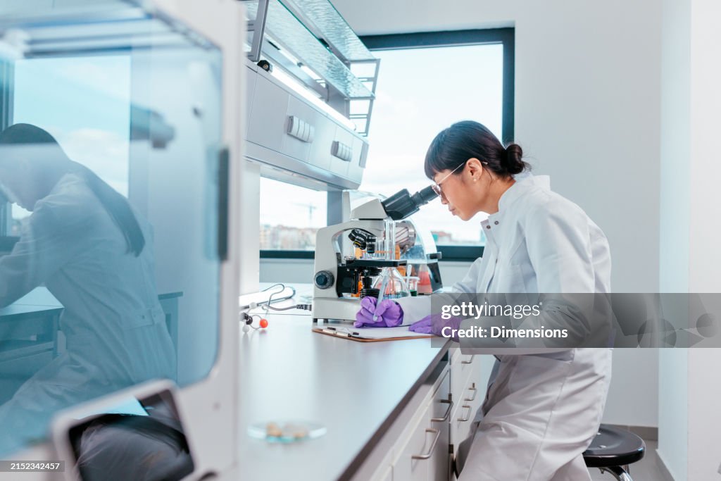 Female scientist looking trough the microscope writing down her discoveries