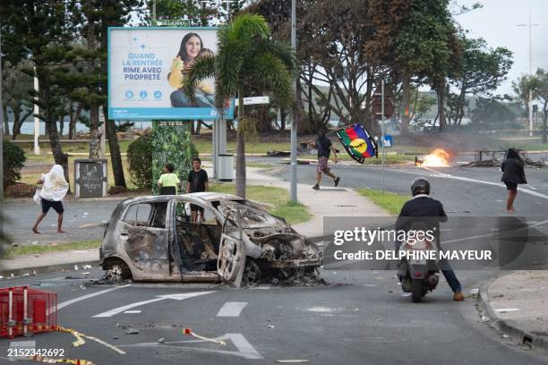 People walk next to a burnt-out car while a man holds a flag of the Socialist Kanak National Liberation Front after a supermarket was looted and...