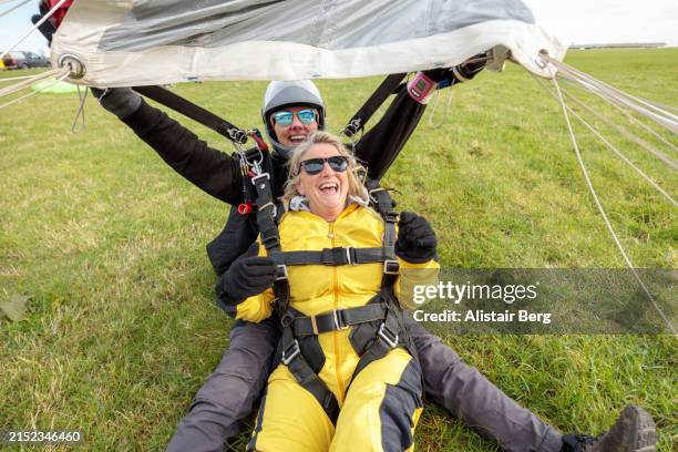 senior woman celebrates as she lands safely with her female instructor after a tandem parachute jump - baby boomer stock pictures, royalty-free photos & images