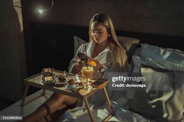 donna che utilizza uno smartphone e mangia un cibo del servizio in camera in una camera d'albergo - servizio in camera foto e immagini stock