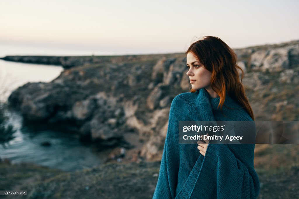 Woman at sunset near the sea in the mountains with a blanket on her shoulders