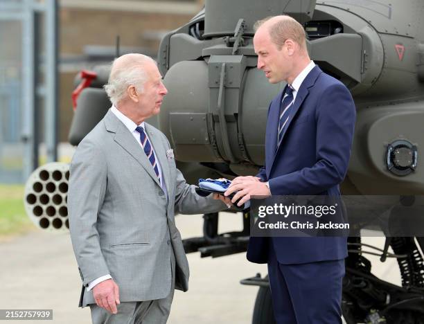 King Charles III hands over the Army Air Corps light blue beret to Prince William, Prince of Wales as they stand in front of an Apache helicopter...