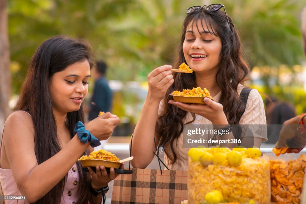 Two young female friends eating indian street food (Bhelpuri) at street market.