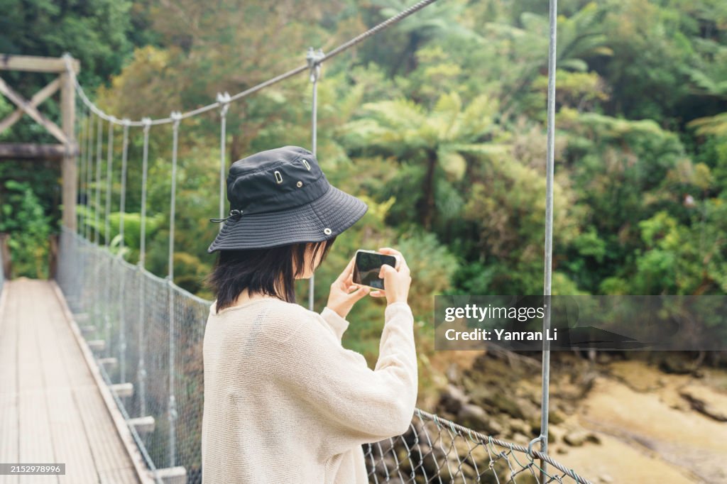 Viajante asiática parando em uma ponte de balanço para tirar fotos usando o smartphone