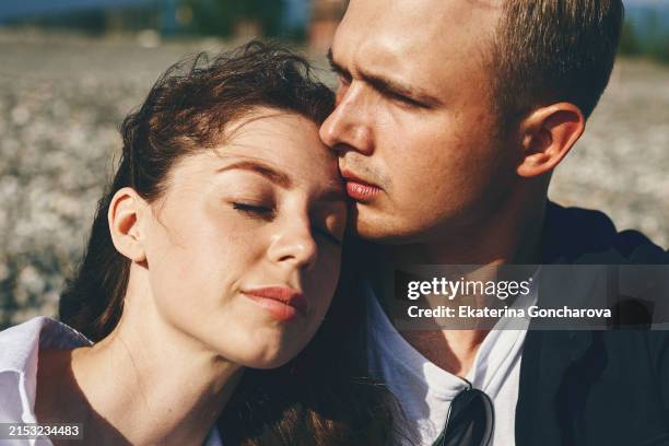 couple sitting close on a beach. woman with closed eyes. man kissing her forehead. casual clothes - stirn stock-fotos und bilder