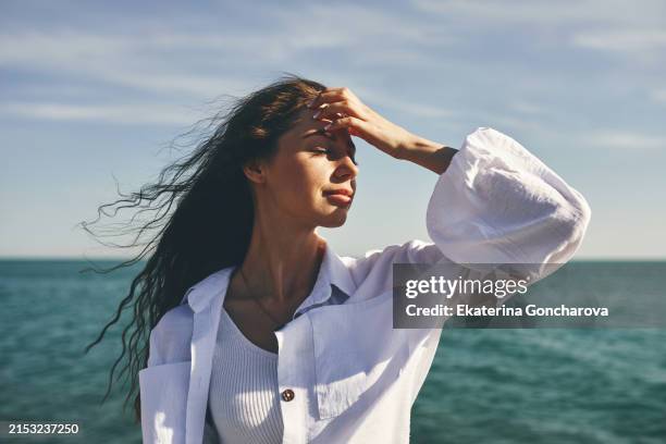 woman standing by the sea with hand on forehead. hair blowing in the wind. clear sky - weathered woman stock pictures, royalty-free photos & images
