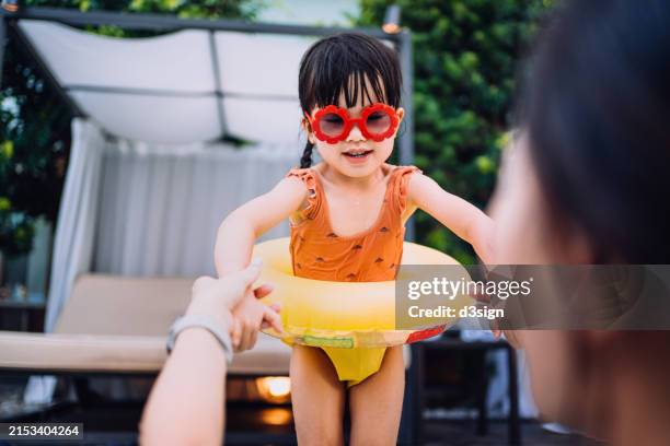 happy little asian girl is having fun in the swimming pool with her mother in summer. mother is holding hands of daughter encouraging her to jump into the pool. enjoying family bonding time on summer vacation. summer fun. travel and vacation concept - young girl jumping into swimming pool stock pictures, royalty-free photos & images