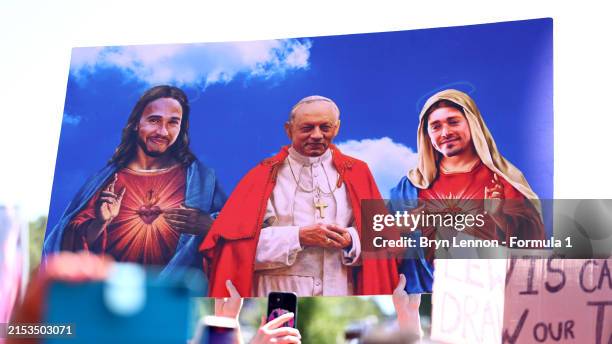 Ferrari fan holds a sign depicting Lewis Hamilton of Great Britain and Mercedes as Jesus, Ferrari Team Principal Frederic Vasseur as the Pope and...