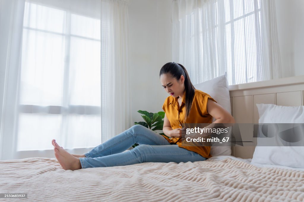 An Asian mid-adult woman in yellow shirt, lying in her bedroom at home, suddenly sits up, experiencing stomach pain, possibly due to uterine fibroids, PMS, or an ulcer.