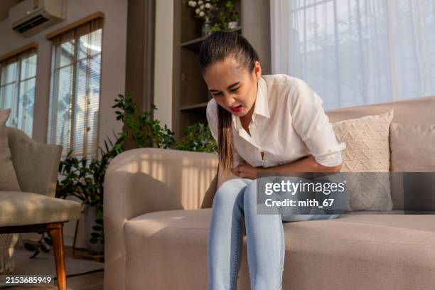 an asian mid-adult woman having a stomachache suddenly while sitting alone on a cozy sofa in a living room at home, due to uterine fibroids, pms, or ulcer - myoma stock pictures, royalty-free photos & images