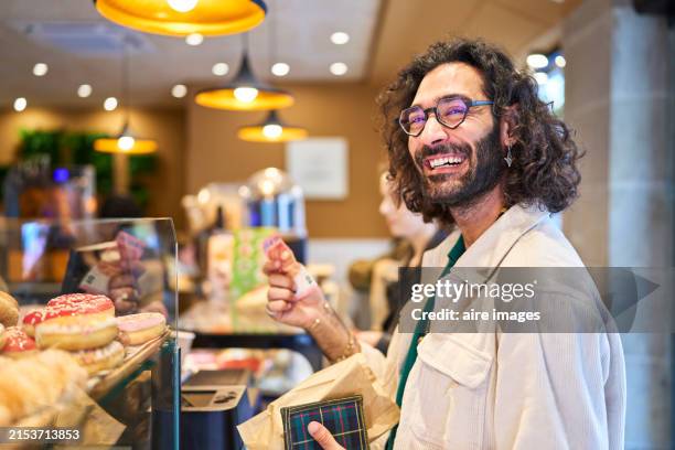 tourist man with glasses and backpack standing in a bakery smiling looking at the camera while paying with a banknote, side view. - grocery store window stock pictures, royalty-free photos & images