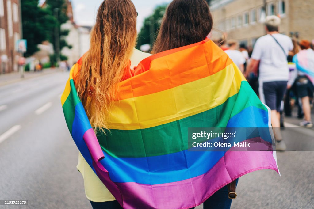 Two female friends wrapped in an LGBT+ flag at the pride parade