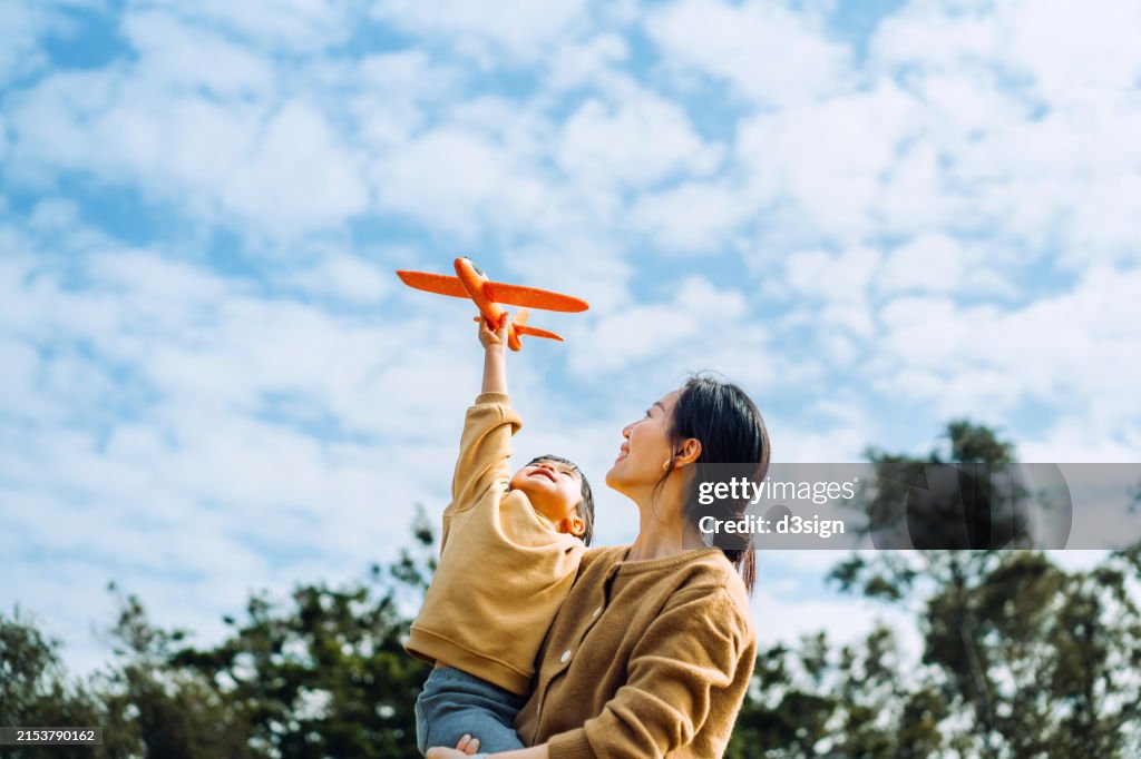 Young Asian mother and lovely little daughter spending time together outdoors, playing with airplane toy and smiling joyfully in park on a lovely sunny day against beautiful blue sky