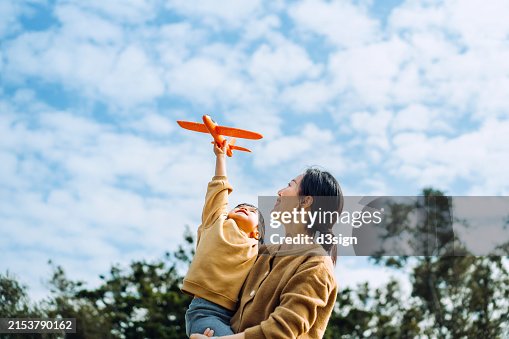 Young Asian mother and lovely little daughter spending time together outdoors, playing with airplane toy and smiling joyfully in park on a lovely sunny day against beautiful blue sky Young Asian mother and lovely little daughter spending time together outdoors, playing with airplane toy and smiling joyfully in park on a lovely sunny day against beautiful blue sky
