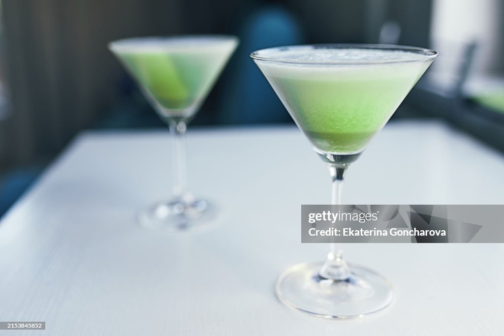 Two green cocktails on a white table. Focus on the front drink with a blurred background.