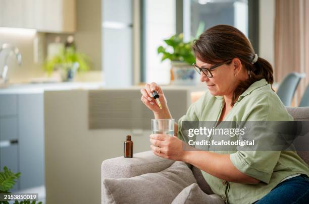 donna matura seduta sul divano usando un contagocce per mettere la medicina in un bicchiere d'acqua - omeopatia foto e immagini stock