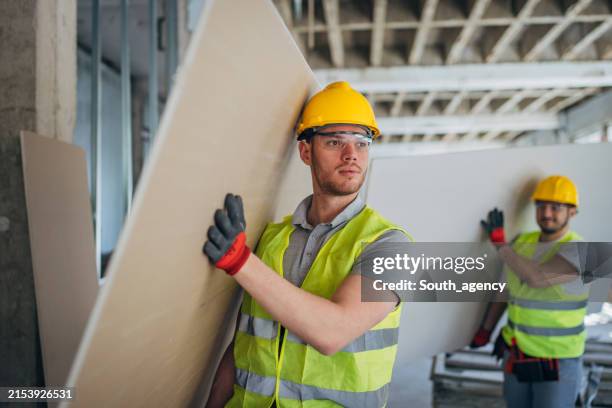 two construction workers in safety gear efficiently transport drywall sheets at a construction site - drywall tools stock pictures, royalty-free photos & images
