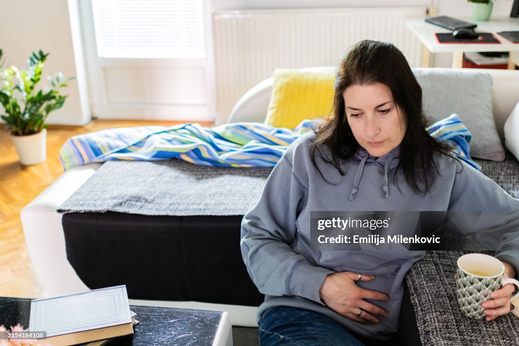 Women with uterine fibroids. Woman is suffering from stomach ache at home. Abdominal pain. Menstrual period, food poisoning, gastritis, diarrhea, feeling unwell sitting in living room.