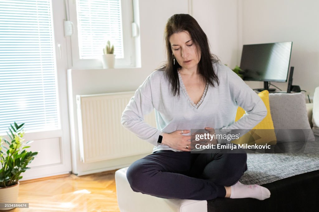 Women with uterine fibroids. Woman is suffering from stomach ache at home. Abdominal pain. Menstrual period, food poisoning, gastritis, diarrhea, feeling unwell sitting in living room.