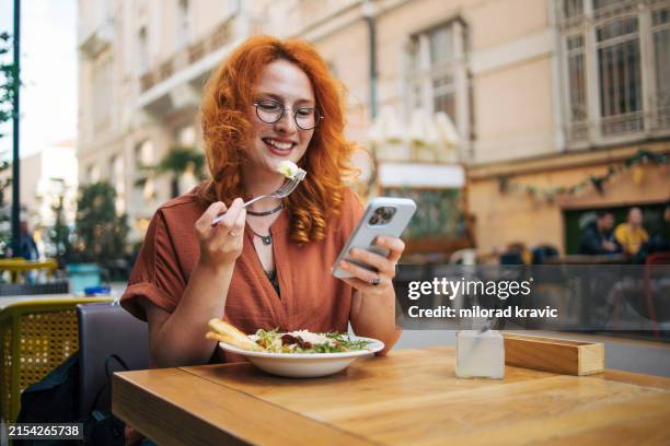 happy young redhead woman eating caesar salad in restaurant, smart phone - caesar salad stock-fotos und bilder