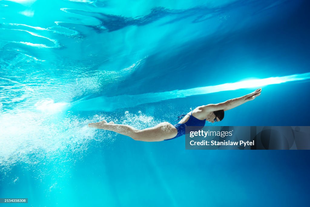 Underwater view on female swimmer in blue swimsuit after the starting jump against transparent blue water