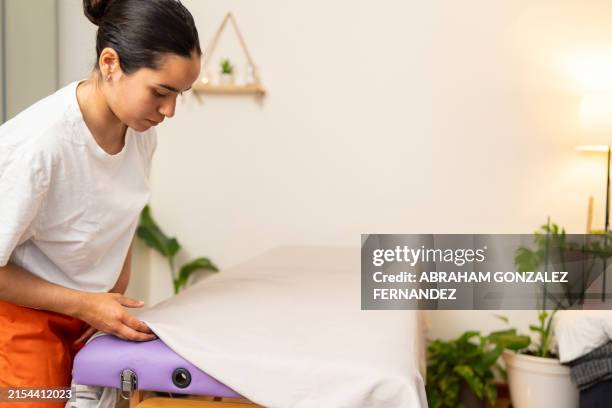 hispanic reiki practitioner sets up a massage table for a wellness session in a tranquil, plantfilled healing space - massage table stock pictures, royalty-free photos & images