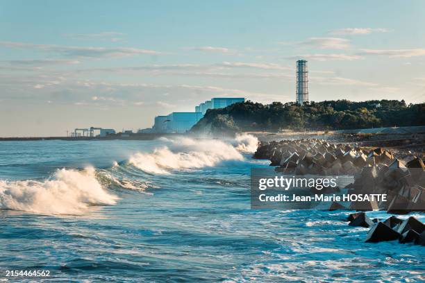 Vagues se projetant sur des blocs de béton sur la plage et à l'arrière plan la centrale nucléaire de Fukushima Daiichi en novembre 2023.