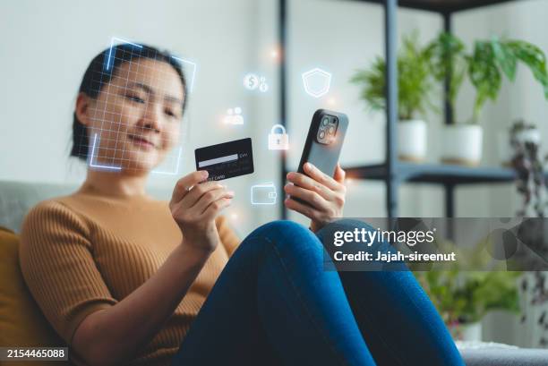 asian woman holding credit card and scanning face id by smart phone for shopping online sitting on sofa in living room at home. - open banking stock pictures, royalty-free photos & images