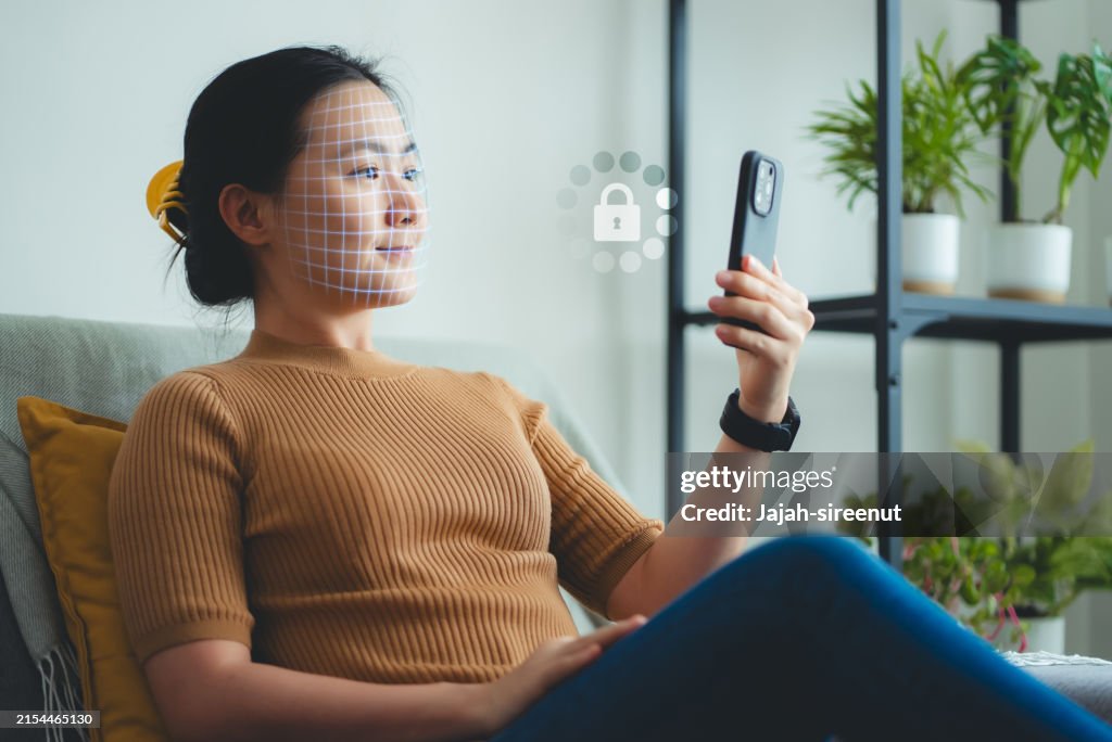 Asian woman scanning face id by smart phone sitting on sofa in living room at home.