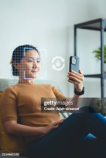 asian woman scanning face id by smart phone sitting on sofa in living room at home. - digitale authenticatie stockfoto's en -beelden