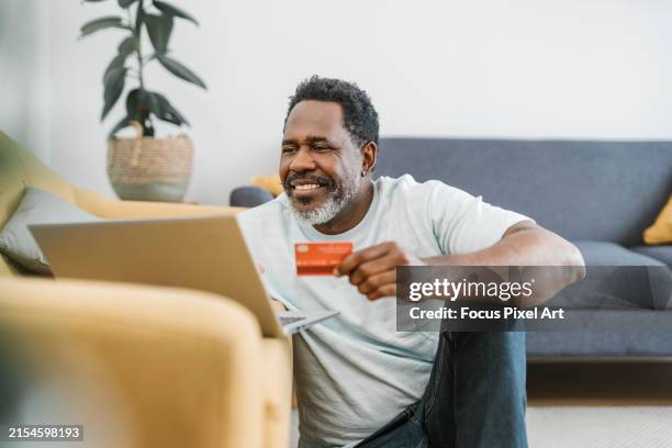 happy mature man sitting in the living room shopping online using a card and laptop. - credit card art stock pictures, royalty-free photos & images