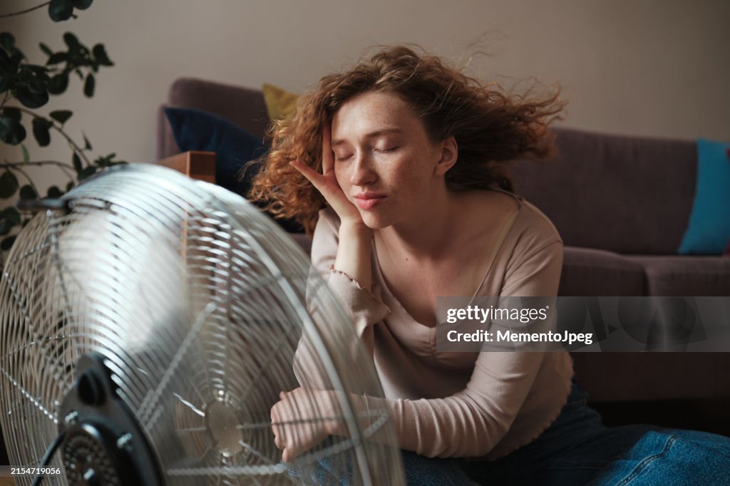 Carefree woman relaxing near electric fan at home, cooling down refreshing during heatwave indoors. Summer heat