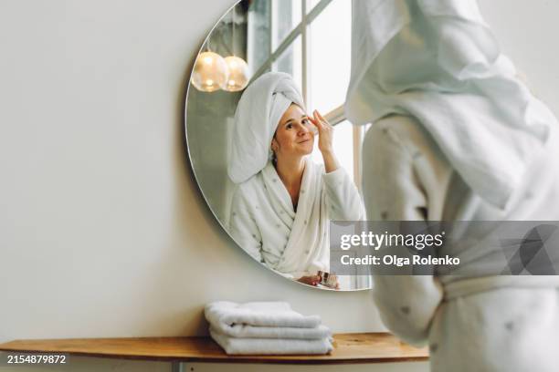 skin brightening routine: smiling woman hydrates skin with moisturizer in mirror reflection - retinol stock pictures, royalty-free photos & images