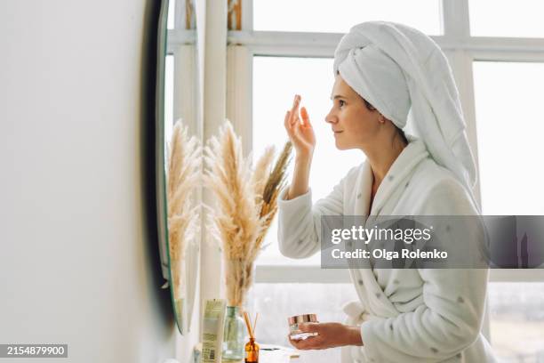 hyperpigmentation care: woman applies balm in bright white bathroom in front of the mirror - hyperpigmentation stockfoto's en -beelden