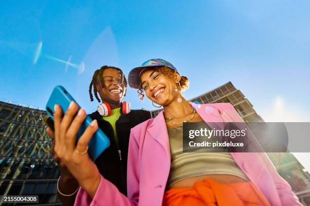 two gen z friends using a smartphone together. low angle shot with modern buildings in the background. social media and friendship concept. - groothoek stockfoto's en -beelden
