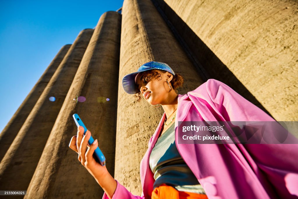 Young African American woman in colorful clothing using a smartphone. Low angle shot with concrete silos in the background. Urban fashion and technology concept.