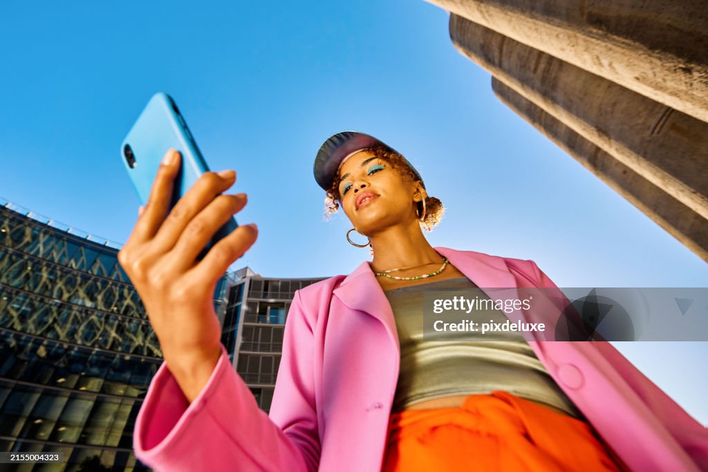 Young African American woman in colorful clothing using a smartphone. Low angle shot with concrete silos in the background. Urban fashion and technology concept.