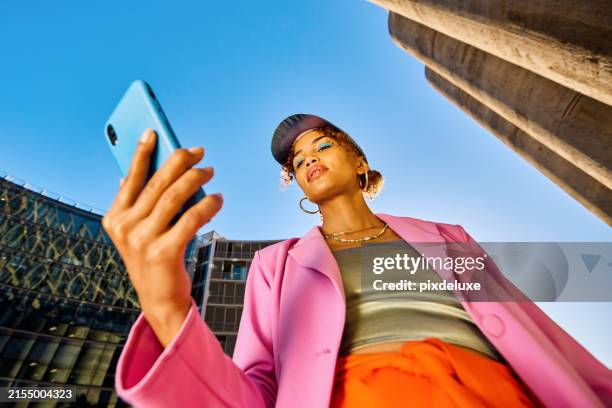 young african american woman in colorful clothing using a smartphone. low angle shot with concrete silos in the background. urban fashion and technology concept. - city colorful photos et images de collection