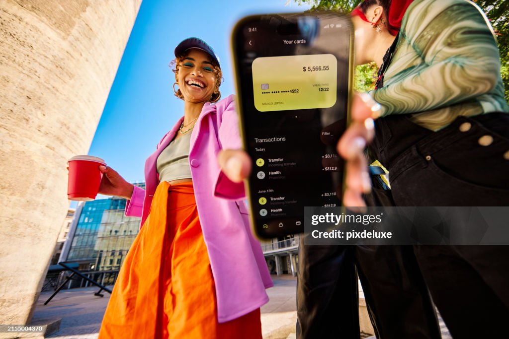 Gen Z adults outdoors with one person showing a smartphone displaying a bank balance and transactions. Urban environment. Low angle shot.