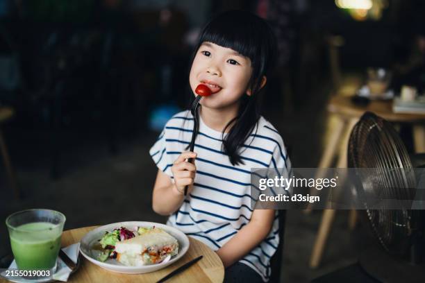 lovely little asian girl eating in cafe, she is enjoying her sandwich and salad lunch set at dining table. having a fun time dining out with her family. people, food and lifestyle - tomate cerise photos et images de collection
