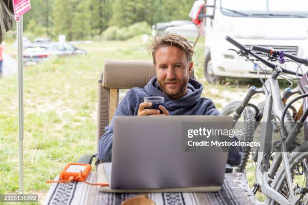 man drinking coffee while working on laptop at campsite - off the grid stockfoto's en -beelden