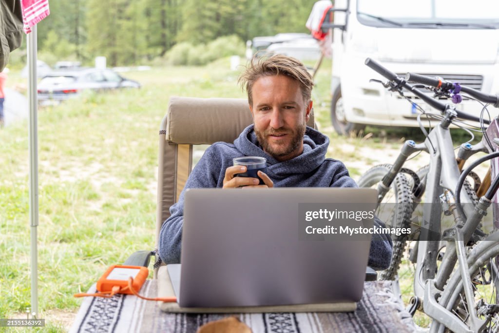 Man Drinking Coffee While Working on Laptop at Campsite