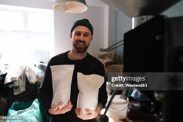 smiling coffee roaster holding packed paper bags of coffee beans in factory - coffee roasting machines stock pictures, royalty-free photos & images