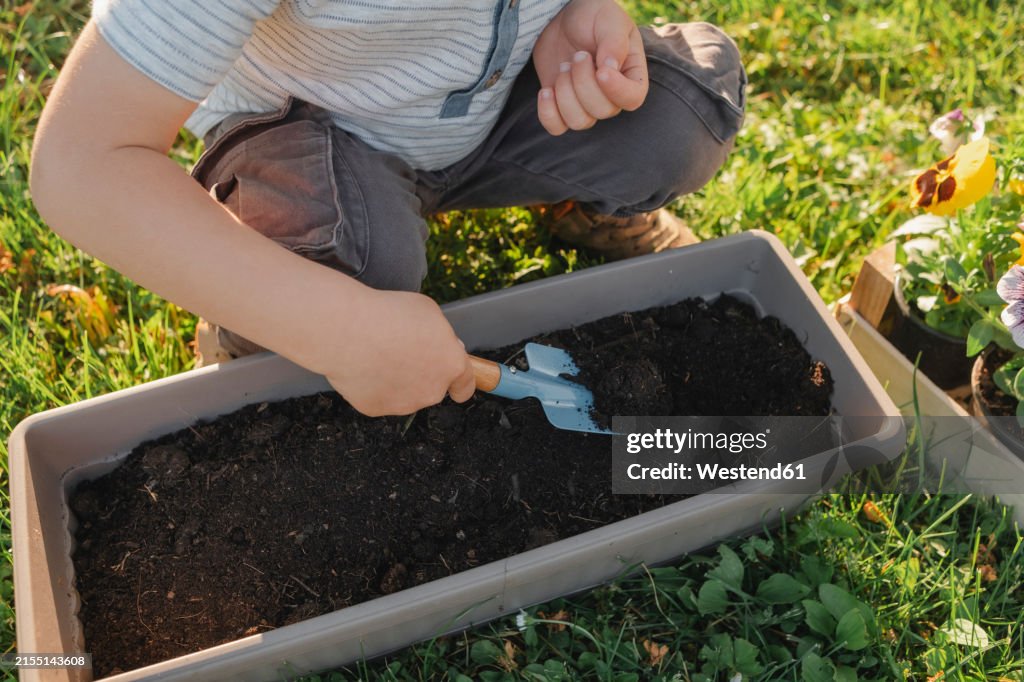 Boy crouching near pot and digging soil using shovel in garden