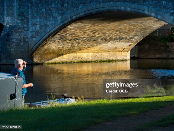 Houseboat moored by Abingdon's medieval stone bridge over the Thames at sunrise, passenger watching.