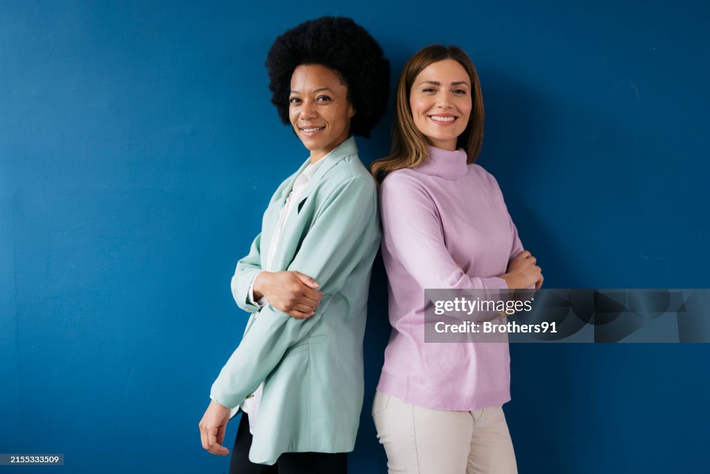 Portrait of two confident businesswomen standing back to back against blue wall