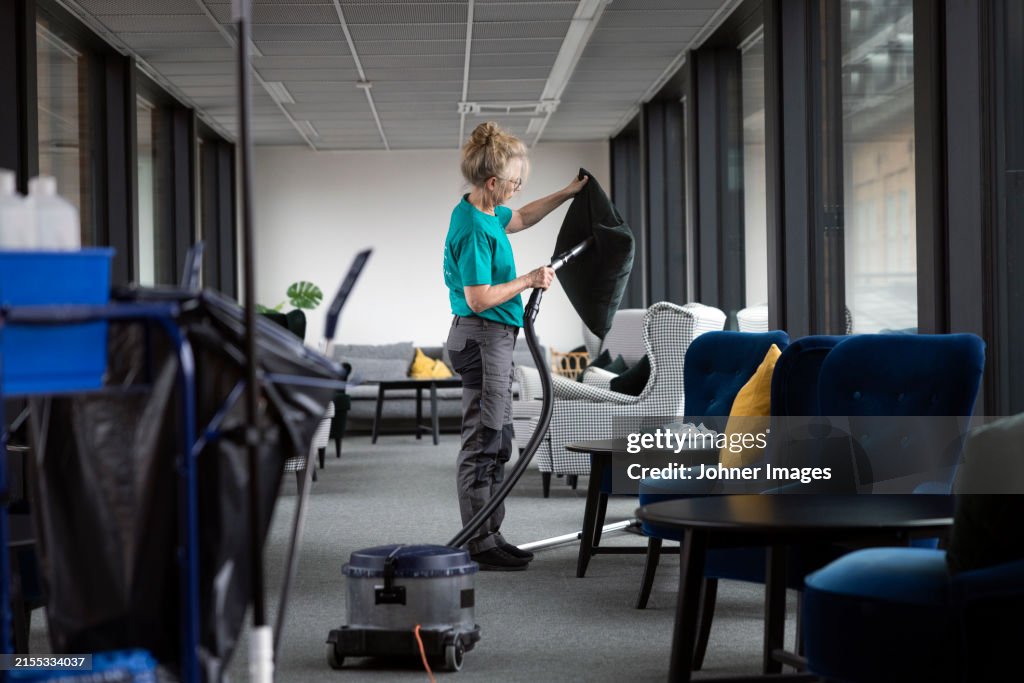 Mature female janitor using vacuum cleaner on cushion while cleaning lobby