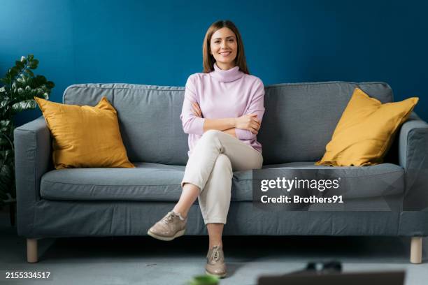 portrait de jeune femme heureuse avec les bras croisés assis sur un canapé gris - assis en tailleur photos et images de collection
