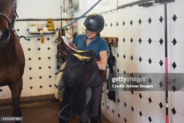 female trainer carrying saddle near horse at paddock - protective sportswear stock pictures, royalty-free photos & images